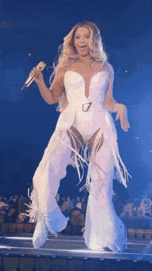 a woman in a white bodysuit and fringe pants is singing into a microphone on a stage