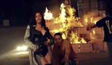 a man wearing a v on his back stands next to a woman in front of a fire