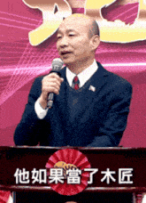 a man in a suit stands at a podium holding a microphone with chinese writing on it