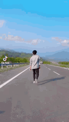 a man is walking down a highway with mountains in the background and a blue sky .
