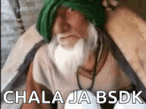 a man with a beard and a green turban is sitting down and talking to the camera .