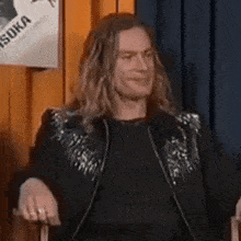 a man with long hair is sitting in a chair wearing a black jacket with sequins on it