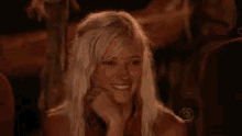 a woman with blonde hair is smiling while sitting in a dark room with her hand on her chin .
