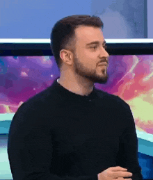 a man with a beard is wearing a black sweater and standing in front of a television .
