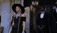 a woman in a black dress and hat is standing in front of a man in a tuxedo .