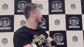 a man in a black shirt is standing in front of a checkered wall with logos on it .