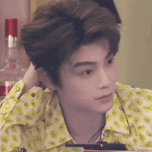 a young man in a yellow and white floral shirt is sitting at a table with his hand in his hair .