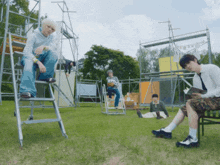 a group of young men are standing in a grassy field with a man sitting on a ladder reading a book