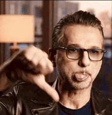 a man wearing glasses and a leather jacket is sticking out his tongue and giving a thumbs down sign .