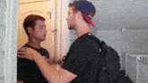 two men are standing next to each other in a room . one of the men is wearing a backpack .