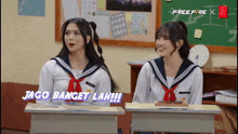 two girls are sitting at desks in a classroom with the words jago banget lah written on the screen behind them