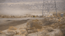 a yellow truck is driving down a dirt road with a white van in the background