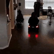 a child is riding a toy car in a living room with a christmas tree in the background .