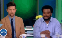 two men are sitting next to each other on a television show . one of the men is wearing a suit and tie .