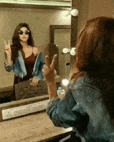 a woman is making a peace sign in front of a mirror while wearing sunglasses and a denim jacket .
