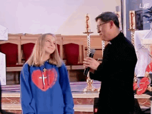 a girl wearing a blue sweatshirt with a heart and cross on it stands next to a priest holding a microphone
