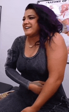 a woman with purple hair is sitting in front of a wall with a poster that says anatomy of .