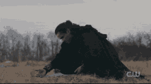 a man in a black cape is kneeling down in a field with a cw logo in the background