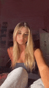 a woman with long blonde hair is sitting on a bed in a room taking a selfie .