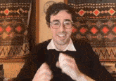 a man wearing glasses and a black sweater is smiling in front of a wall with a patterned curtain .