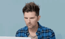 a man in a plaid shirt is sitting in front of a laptop computer and making a funny face .