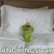 kermit the frog is sitting on a bed with white sheets and pillows .