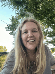 a woman with long blonde hair smiles in front of trees
