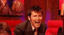 a man in a suit and tie is laughing while sitting on a red couch ..