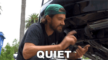 a man wearing a green hat and a black shirt is working on a car and the word quiet is next to him