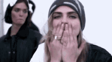 a woman in a beanie is covering her mouth with her hands while another woman looks on .