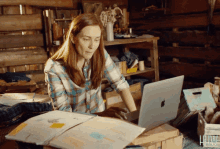 a woman in a plaid shirt is typing on an apple laptop in a cluttered room