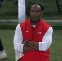 a man wearing headphones and a red vest is sitting on a bench with his arms crossed on a football field .