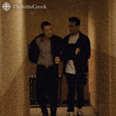 two men are walking down a hallway with #schittscreek written on the wall behind them