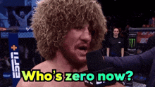 a man with curly hair is talking into a microphone with the words who 's zero now below him