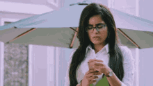 a woman wearing glasses is standing under an umbrella while looking at her phone .