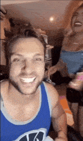 a man with a beard wearing a blue tank top is smiling while a woman looks on .