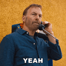 a man in a blue shirt is talking on a payphone and the word yeah is on the bottom right