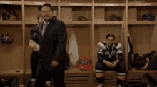a man in a suit is standing in a locker room with a hockey player sitting on a bench