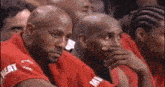 a group of basketball players are sitting in the stands watching a game and one of them is covering his mouth with his hand .