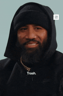 a man with a beard is wearing a black hoodie and a black beanie and says trash