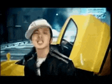 a man wearing a hat is standing next to a yellow car with the hood open .