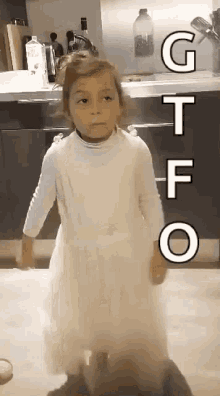 a little girl in a white dress is standing in a kitchen with the letters gtfo written on the wall behind her .