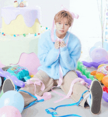 a boy in a blue sweater sits on the floor with balloons and a birthday cake in the background