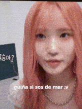 a girl with pink hair is wearing a necklace and looking at the camera while talking in spanish .