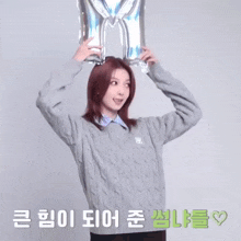 a woman in a grey sweater is standing with her arms in the air with korean writing behind her