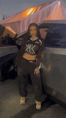 a woman in a crop top with the letter k on it stands in front of a car