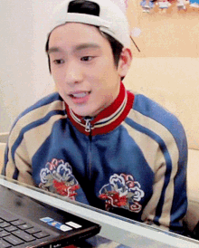 a young man wearing a baseball cap and a blue jacket sits in front of a laptop computer