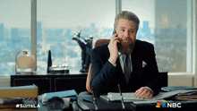 a man in a suit is sitting at a desk talking on his cell phone