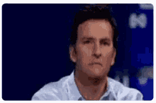 a man in a white shirt is sitting in front of a blue background and making a funny face .