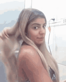 a woman with long blonde hair is holding her hair in a bathroom and looking at the camera .